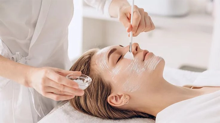 Esthetician applying a hydrating cream facial mask onto a woman's face using a fan brush during a spa treatment.